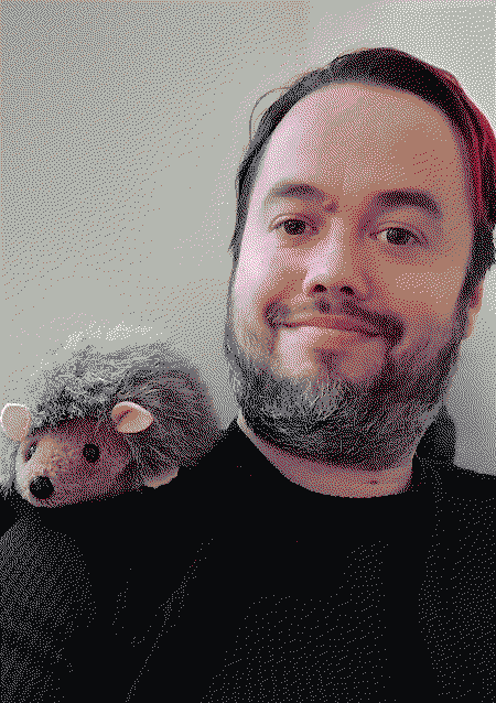 A person with short dark hair and a red stripe through it smiling with a plush hedgehog on his shoulder